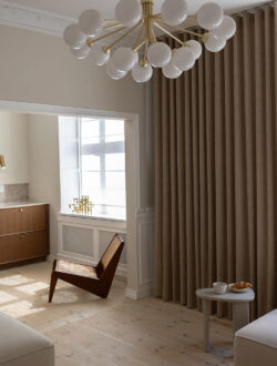 Luxaflex stofgardiner i en mørk beige, der hænger i en stue, hvor der også er en lænestol.